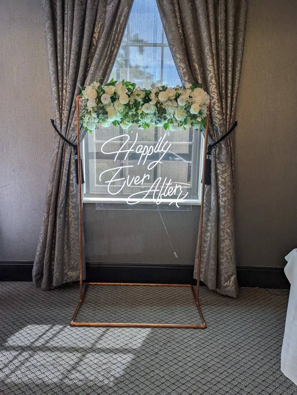 Neon Happily Ever After sign with white floral garland on a copper stand beside draped curtains